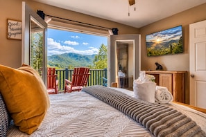 Een slaapkamer met een groot bed, een balkon met stoelen, een schilderij en uitzicht op bergen.