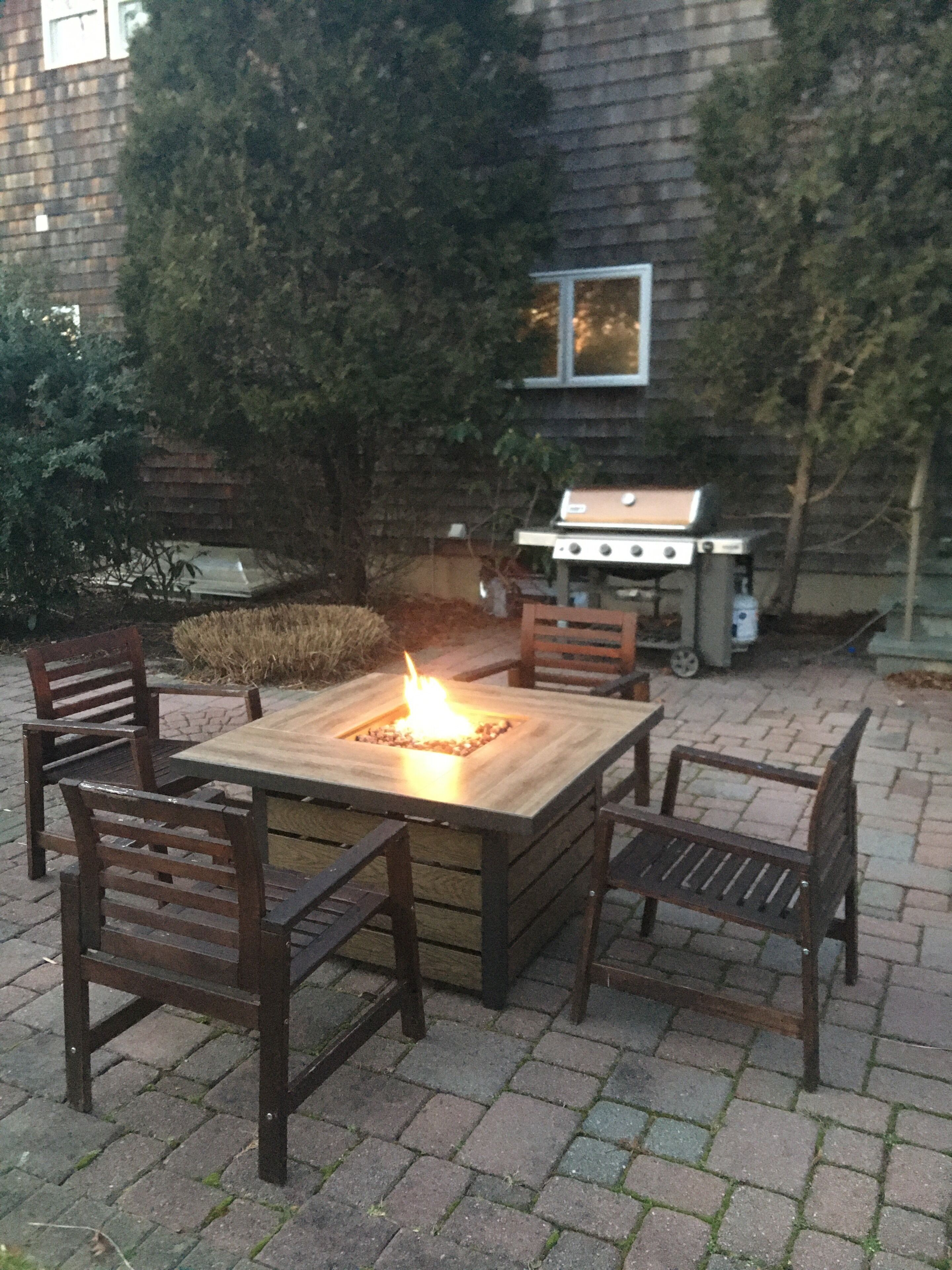 An outdoor patio with a square fire pit table surrounded by wooden chairs. A barbecue grill is visible in the background.