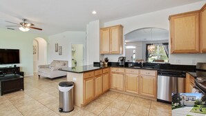 A modern kitchen with wooden cabinets, a black countertop, stainless steel appliances, and a dining area with a sofa and TV. - 6BR Family Retreat Near Disney | Pool + Arcade + Resort Access (Kissimmee)