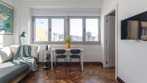 Una sala de estar moderna con un sofá, una mesa de comedor con sillas, una planta en maceta y un ventanal con vistas a la ciudad.