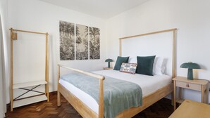 Una cama individual con estructura de madera, una manta verde y dos almohadas. Hay una mesita de noche con una lámpara y una pequeña estantería. El suelo es de madera con patrón espiga y las paredes son blancas.