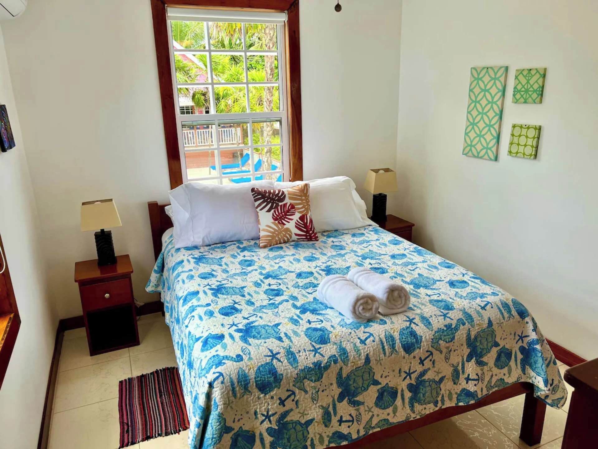 Un lit bien fait, recouvert d’une couvre-lit à motifs bleus et blancs, avec deux oreillers, une serviette enroulée et une table de chevet surmontée d’une lampe.