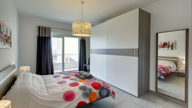 Une chambre à coucher moderne avec un lit, un grand miroir, un luminaire au plafond et une vue sur le balcon extérieur.