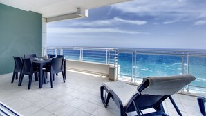 A balcony with a table and chairs, a cushioned lounge chair, and a view of the ocean.