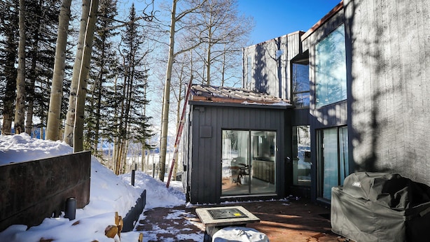 Eine moderne Holzhütte mit Glastür, die auf eine Terrasse führt, umgeben von Schnee und Bäumen.