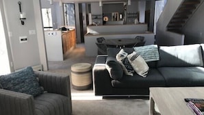 Ein modernes Wohnzimmer mit einem grauen Sofa, einem gestreiften Sessel und einem Couchtisch. Im Hintergrund ist eine Essecke mit Tisch und Stühlen zu sehen.