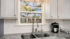 A kitchen with white cabinets, a double sink, a window with a view of trees, and a coffee maker on the counter. - Bella Manor Lavender Suite | King Bed | Walk to Main (Fredericksburg)