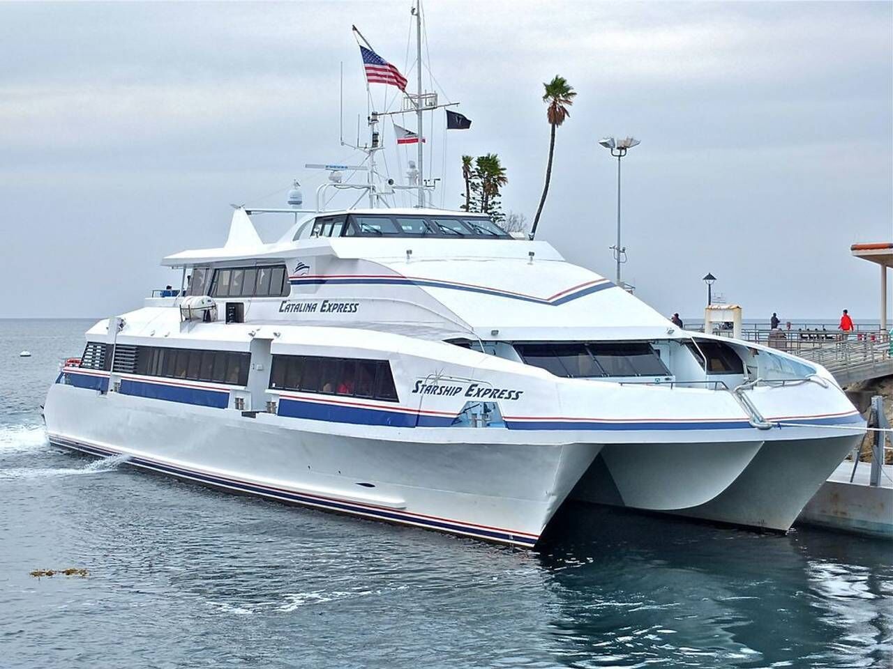 Un yate blanco grande con la marca 'CATHALINA EXPRESS' y 'STARSHIP EXPRESS', atracado en un muelle con un cielo despejado y palmeras al fondo.