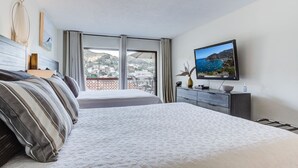 A hotel room with two beds, a TV, a dresser, and a balcony with a view of the sea and buildings.