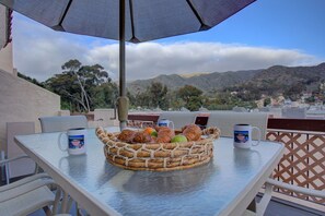 Una mesa con una cesta tejida que contiene pasteles y frutas, dos tazas y vistas a un pueblo costero con montañas al fondo.