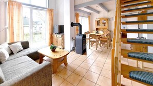 Una sala de estar acogedora con un sofá, una mesa de madera y un televisor. Se ve una escalera con barandilla de madera y una zona de cocina con estufa y armarios.