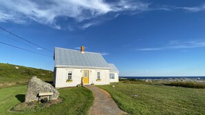 Une petite maison blanche avec un toit gris, une porte jaune et un petit porche. Un grand rocher sur lequel est fixée une pancarte indiquant « Bienvenue » se trouve devant.
