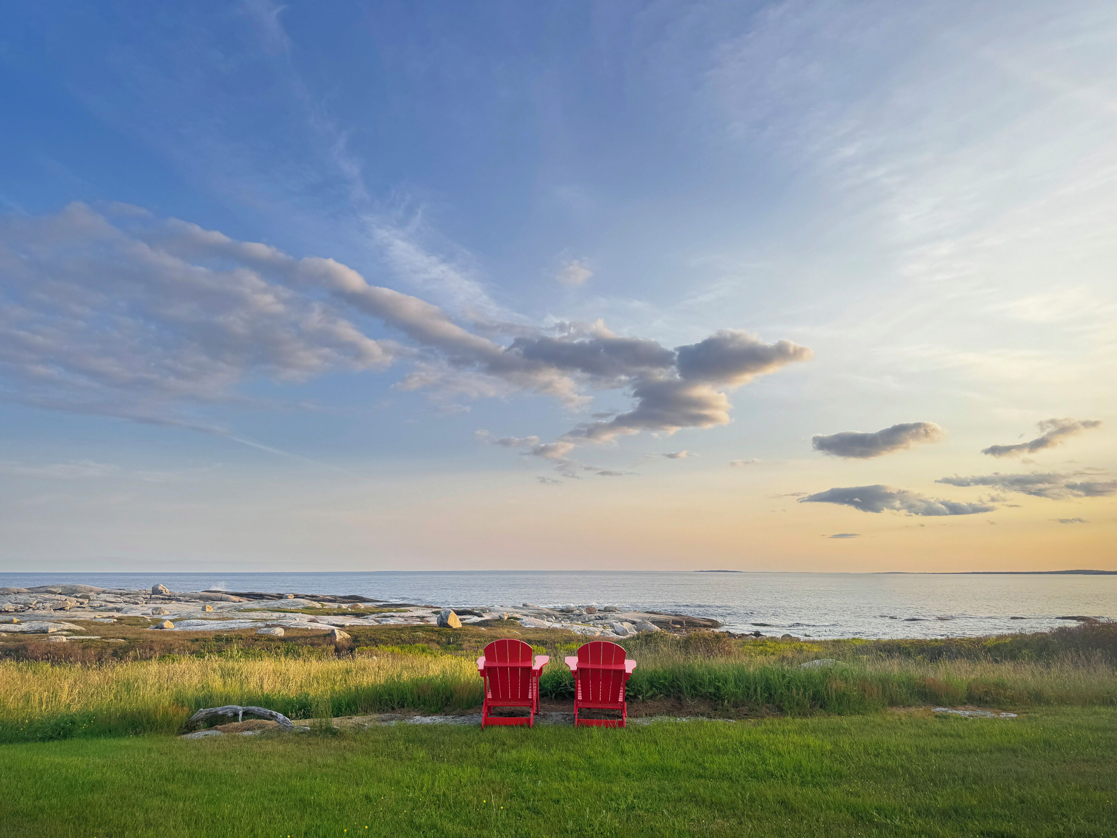 Deux chaises Adirondack rouges sont placées face à une côte rocheuse, offrant une vue sur l’océan et un ciel dégagé.