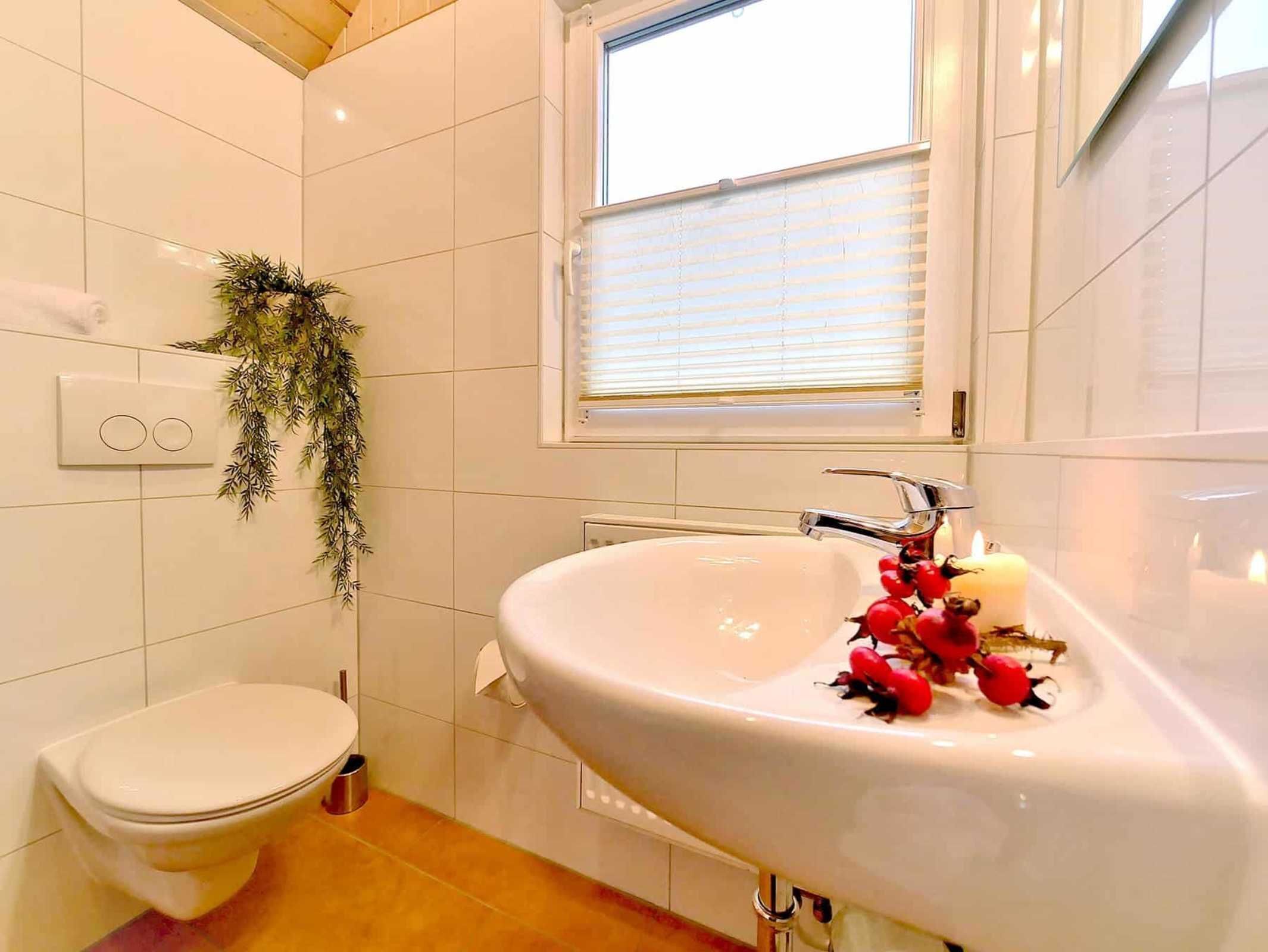 Une salle de bain avec des toilettes blanches, un lavabo avec un miroir et une fenêtre avec des stores.