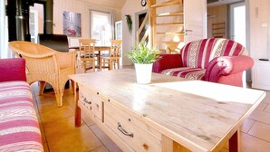 Ein gemütliches Wohnzimmer mit einem Holztisch, Korbstühlen und einem roten Sofa. Eine Treppe führt auf eine erhöhte Ebene.