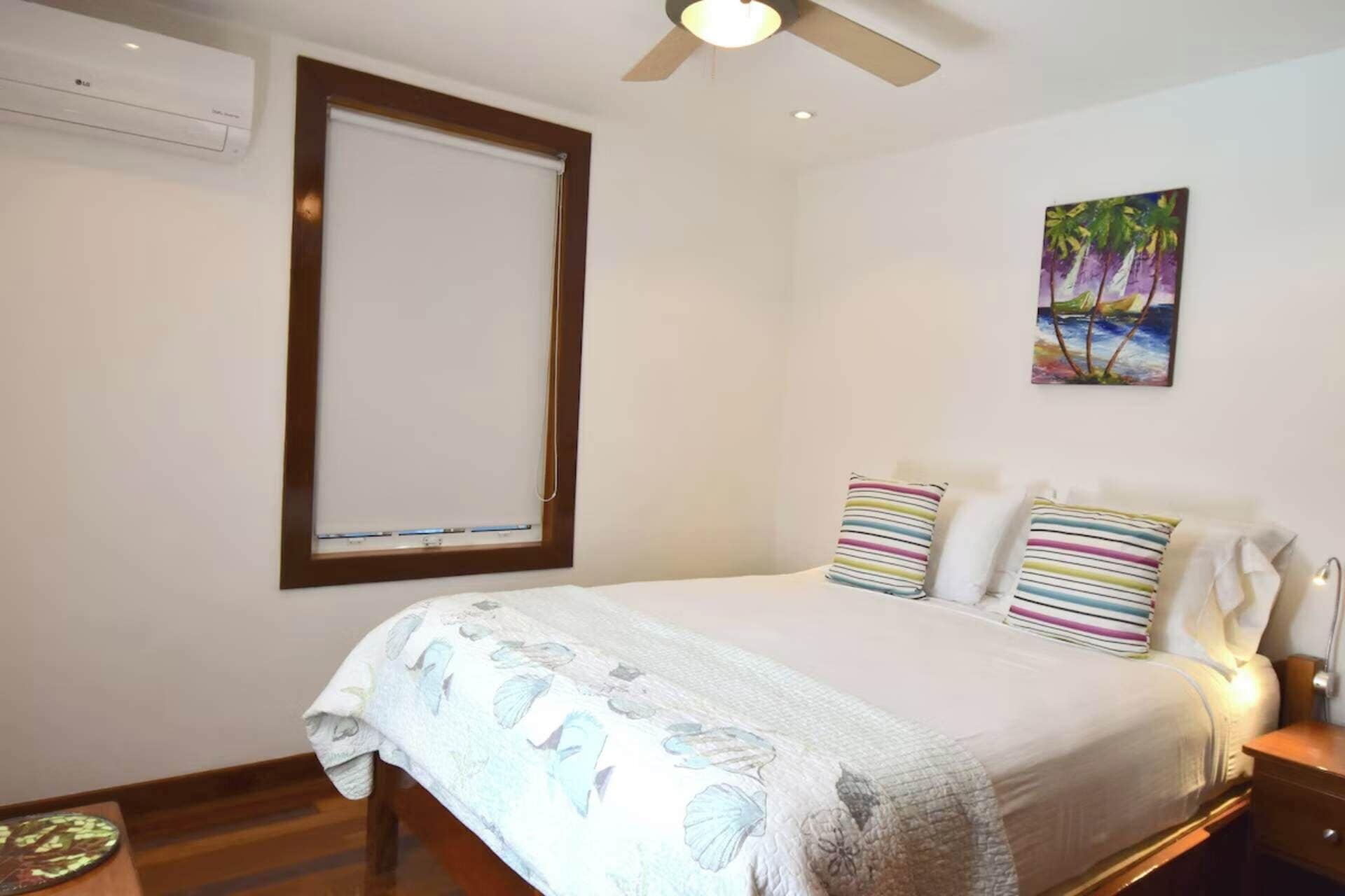 A bedroom with a bed, a ceiling fan, a window with blinds, a painting, and a bedside table.