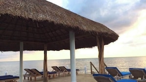 Een terras aan het strand met een houten dek en parasols met rieten dak, met uitzicht op de zee.