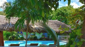 Un área de piscina con estructuras de techo de paja, sillas de descanso y exuberante vegetación tropical.