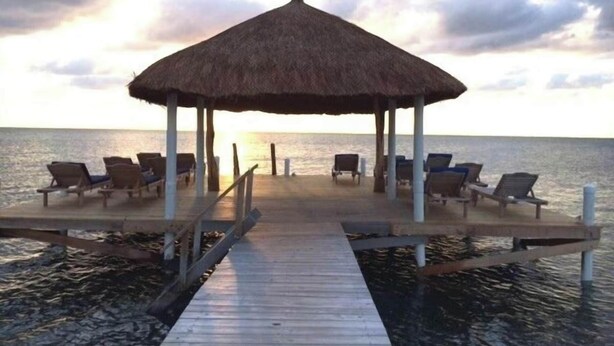 Un muelle de madera que conduce a un pabellón con techo de paja y sillones de descanso, con vista al mar.
