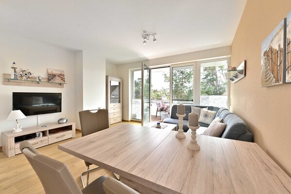 A modern living room with a wooden dining table, a flat-screen TV, and a sofa with cushions.