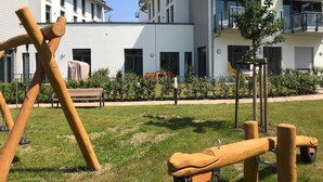 Un parque infantil con columpios y un neumático colgante, rodeado de un césped cuidado y un edificio con varias ventanas y balcones.