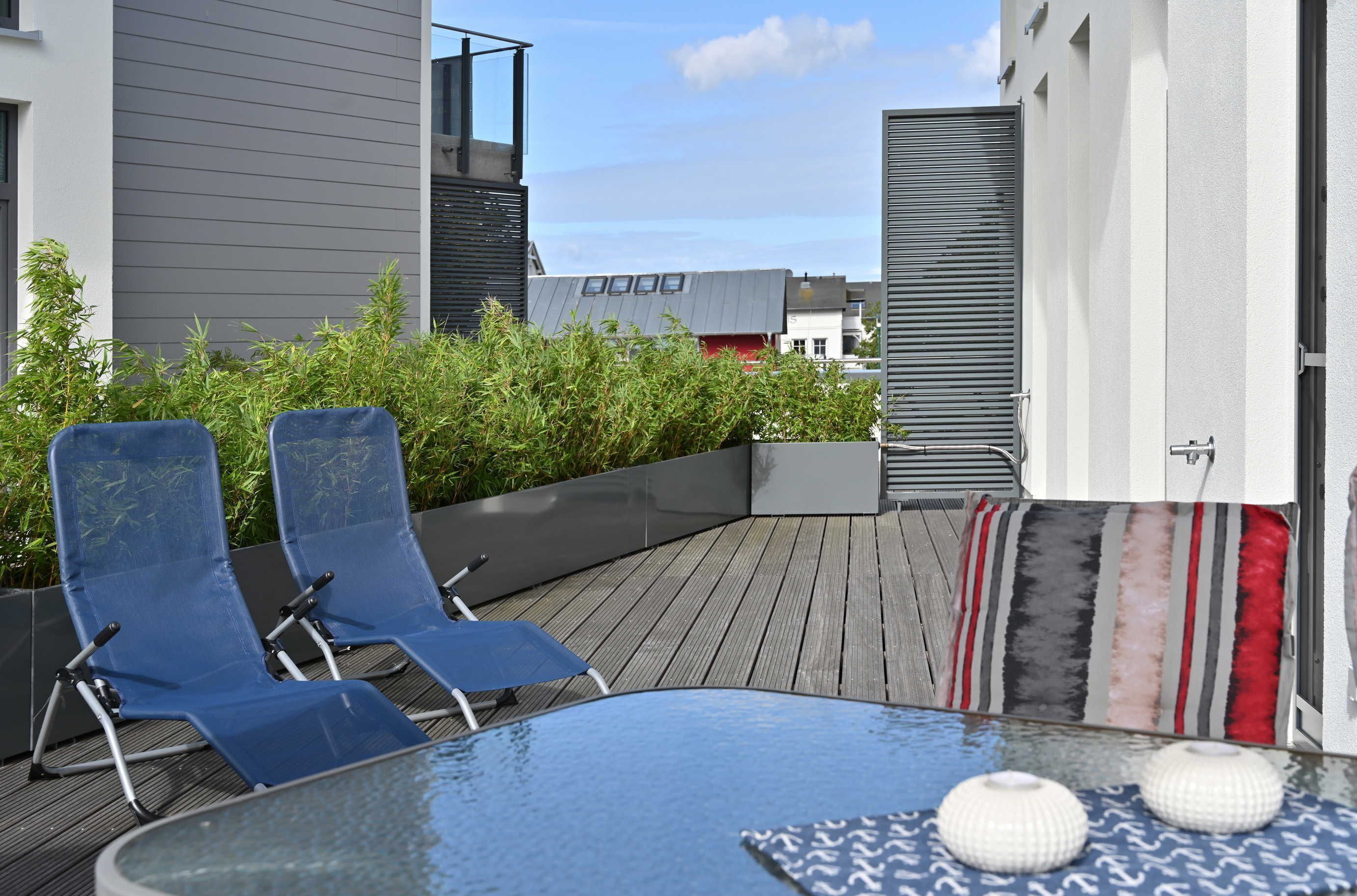 A rooftop terrace with two blue lounge chairs, a glass table, and a decorative pillow.