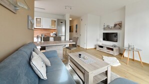 Una sala de estar moderna con sofá, mesa de centro y zona de televisión. Se observa una cocina al fondo, con armarios y un refrigerador.