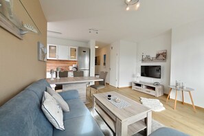 Un salon moderne avec un canapé, une table basse et un espace dédié à la télévision. On aperçoit, au fond, une cuisine équipée de placards et d’un réfrigérateur.