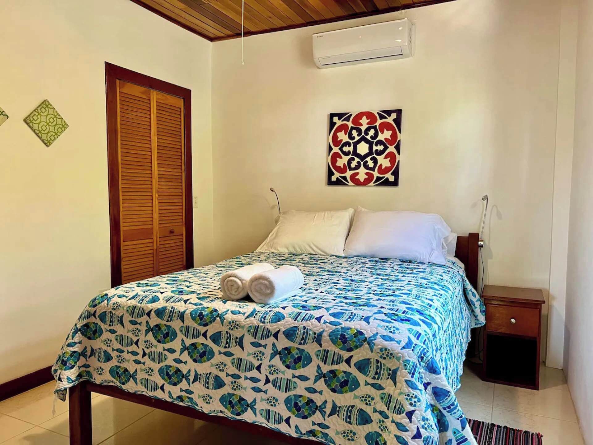 A bedroom with a bed, a nightstand, an air conditioning unit, and a decorative wall piece.