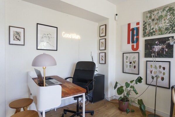 A modern office space with a desk, chair, and laptop. There are framed botanical prints on the walls and a neon sign that reads "Good Vibes."