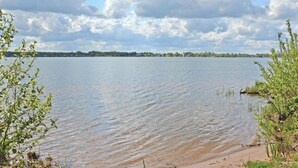 Ein ruhiger See mit sandigem Ufer, umgeben von viel Grün und einer fernen Uferlinie mit Gebäuden.