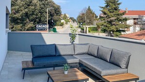Een modern buitenzitgedeelte met een bank en twee bijzettafeltjes, een potplant en een bord met 'CACADU CAFES' op de achtergrond.