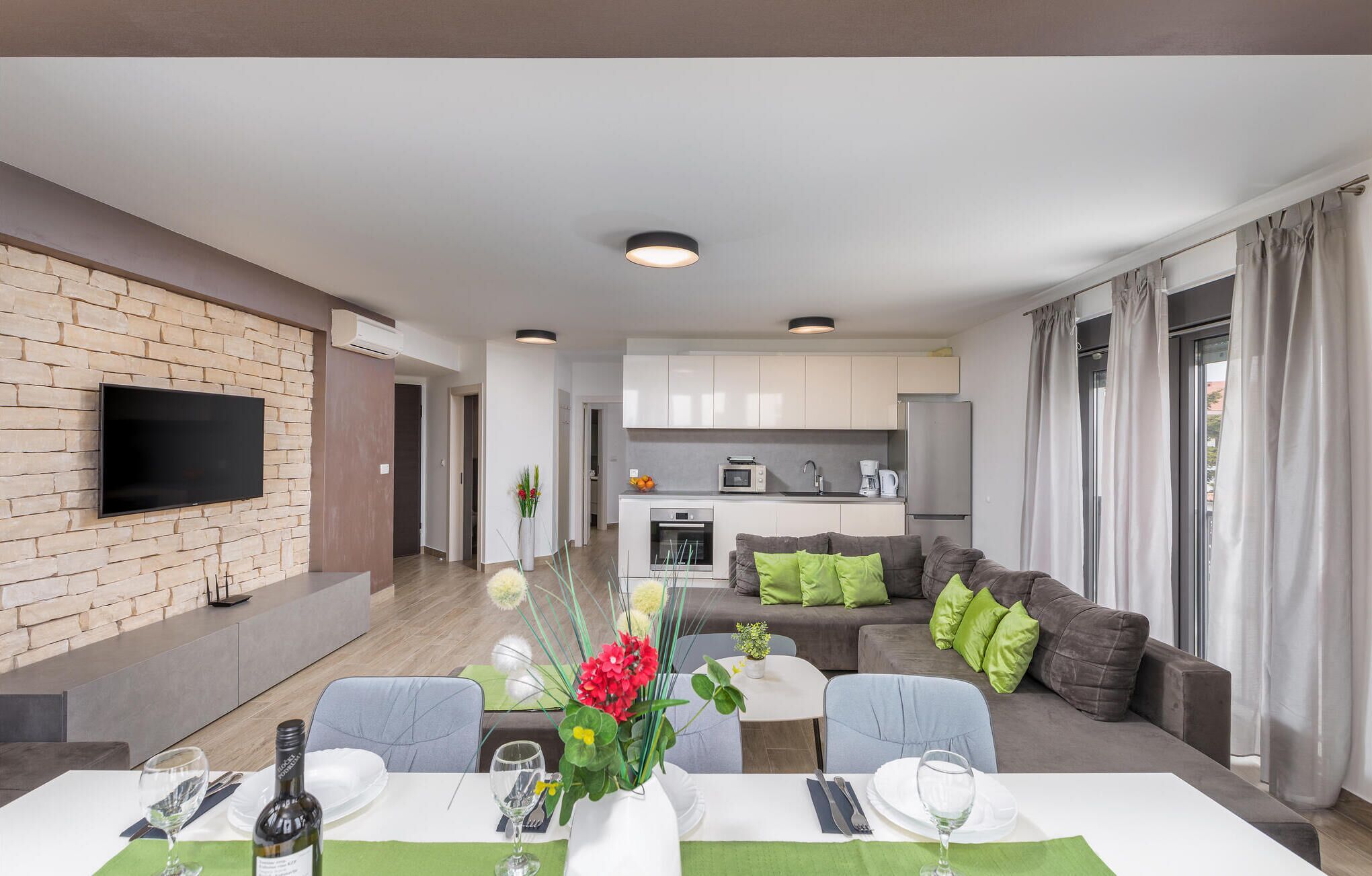 Um apartamento moderno com área de jantar, sala de estar com sofá e almofadas verdes, cozinha com armários brancos e uma TV de tela plana montada em uma parede de tijolos.