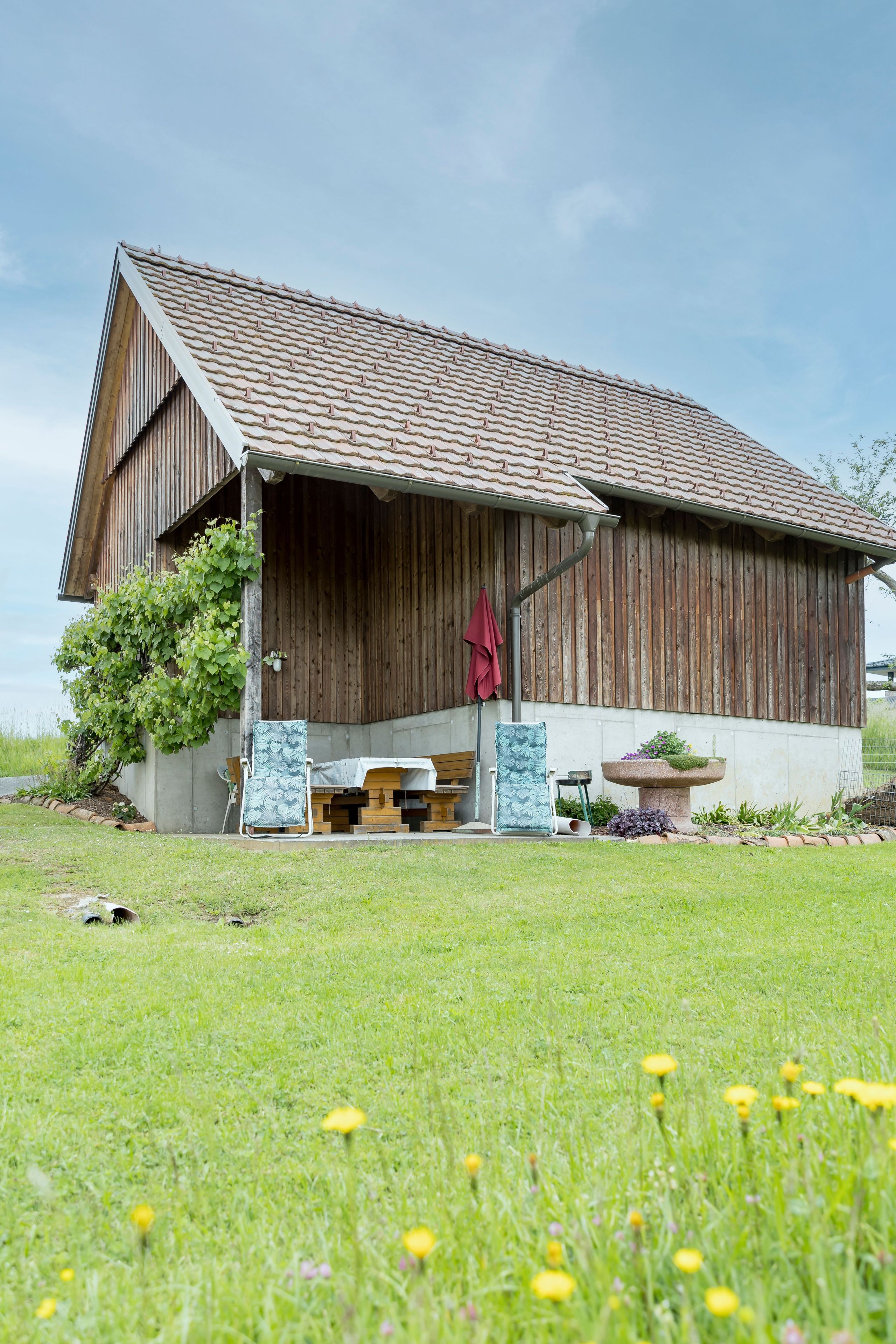 Une grange en bois avec un toit en pente, une véranda et un espace jardin agrémenté d’un banc et d’un jardinière.