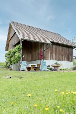A wooden barn with a sloped roof, a covered porch, and a garden area with a bench and a planter. - Vacation cottage - Ermi/Pracher (Petzelsdorf in der Weststeiermark)