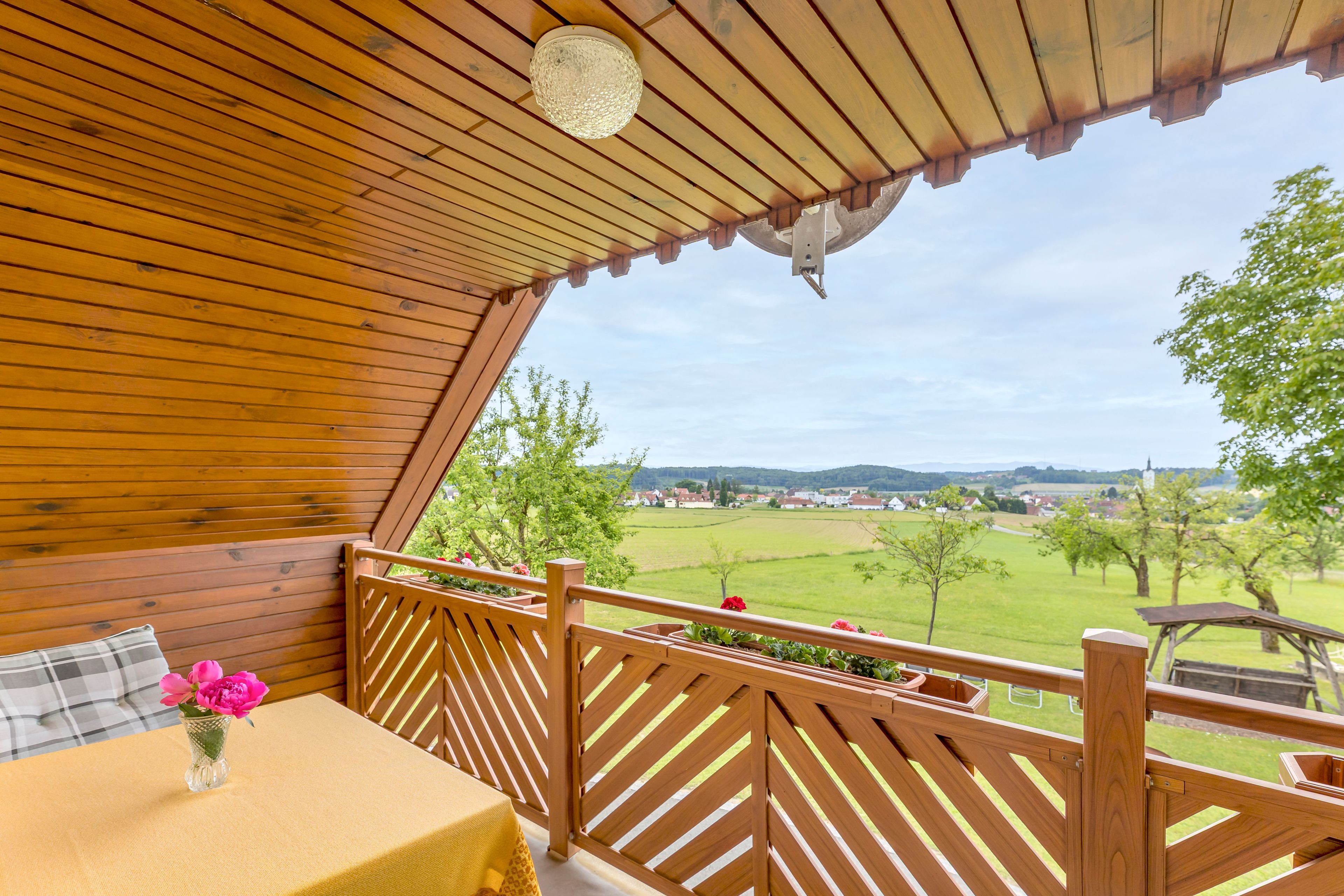 Una terraza de madera con una mesa y un jarrón de flores, con vistas a un paisaje verde.