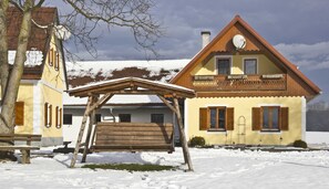 Un chalet a due piani con un balcone in legno, un'altalena di legno e un tetto imbiancato dalla neve.