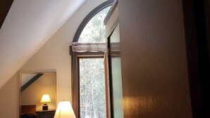Une chambre avec un plafond en pente, une fenêtre donnant sur les arbres, une commode et une lampe.
