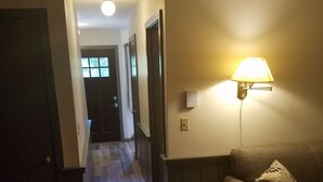 Un couloir au plancher de bois menant à une porte, avec une lampe fixée au mur et un canapé agrémenté d’un coussin.