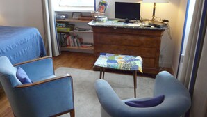 Une chambre à coucher avec un lit, deux chaises, un bureau avec un ordinateur, des étagères et une fenêtre.