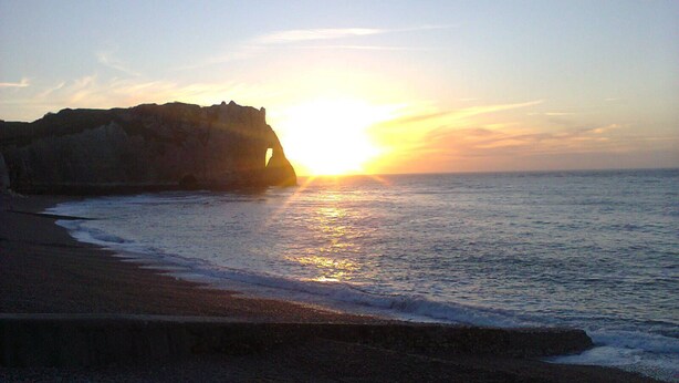Une plage avec un promontoire rocheux et le soleil se couchant sur l’océan.