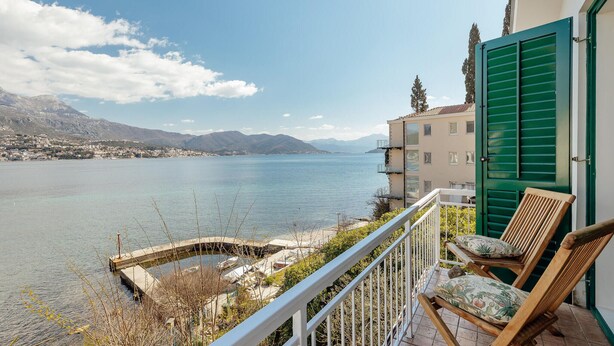 A balcony with a view of a lake, a chair, and a building with green shutters. - Stunning home in Njivice with WiFi (Njivice)