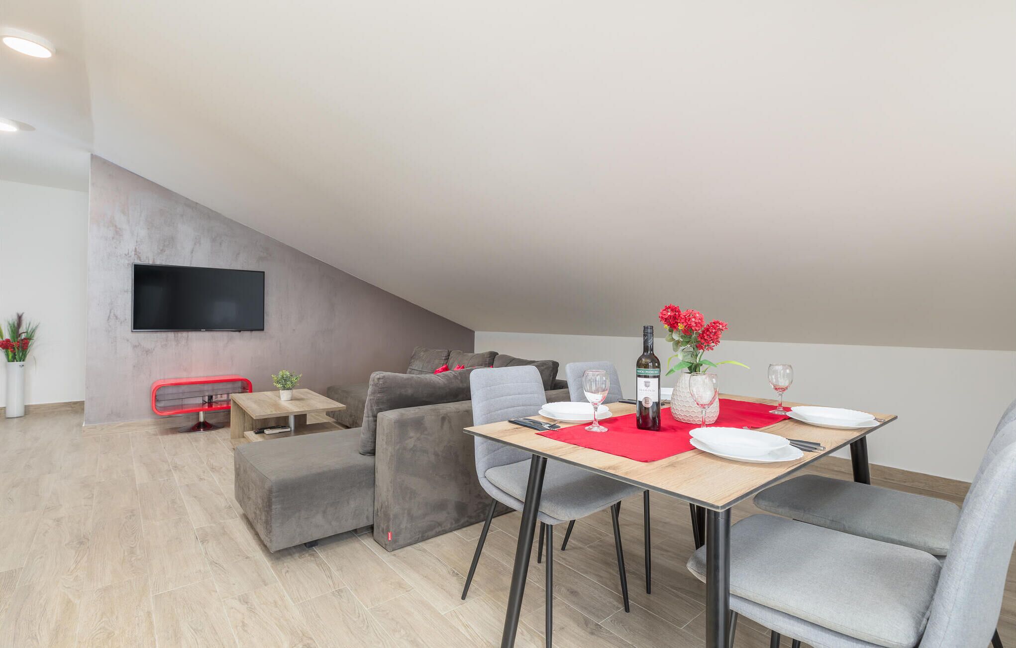 A modern living room with a dining area, grey sofas, a flat-screen TV, and a red tablecloth.