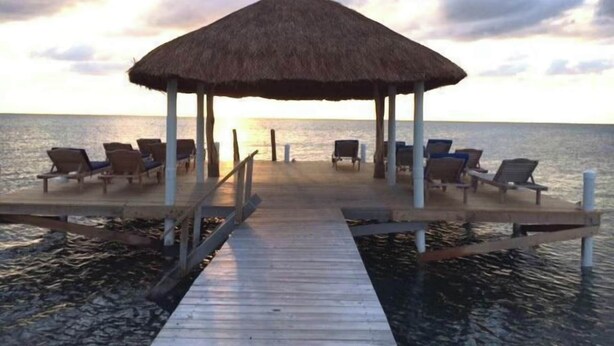 Un muelle de madera que conduce a un pabellón con techo de paja y sillones de descanso.