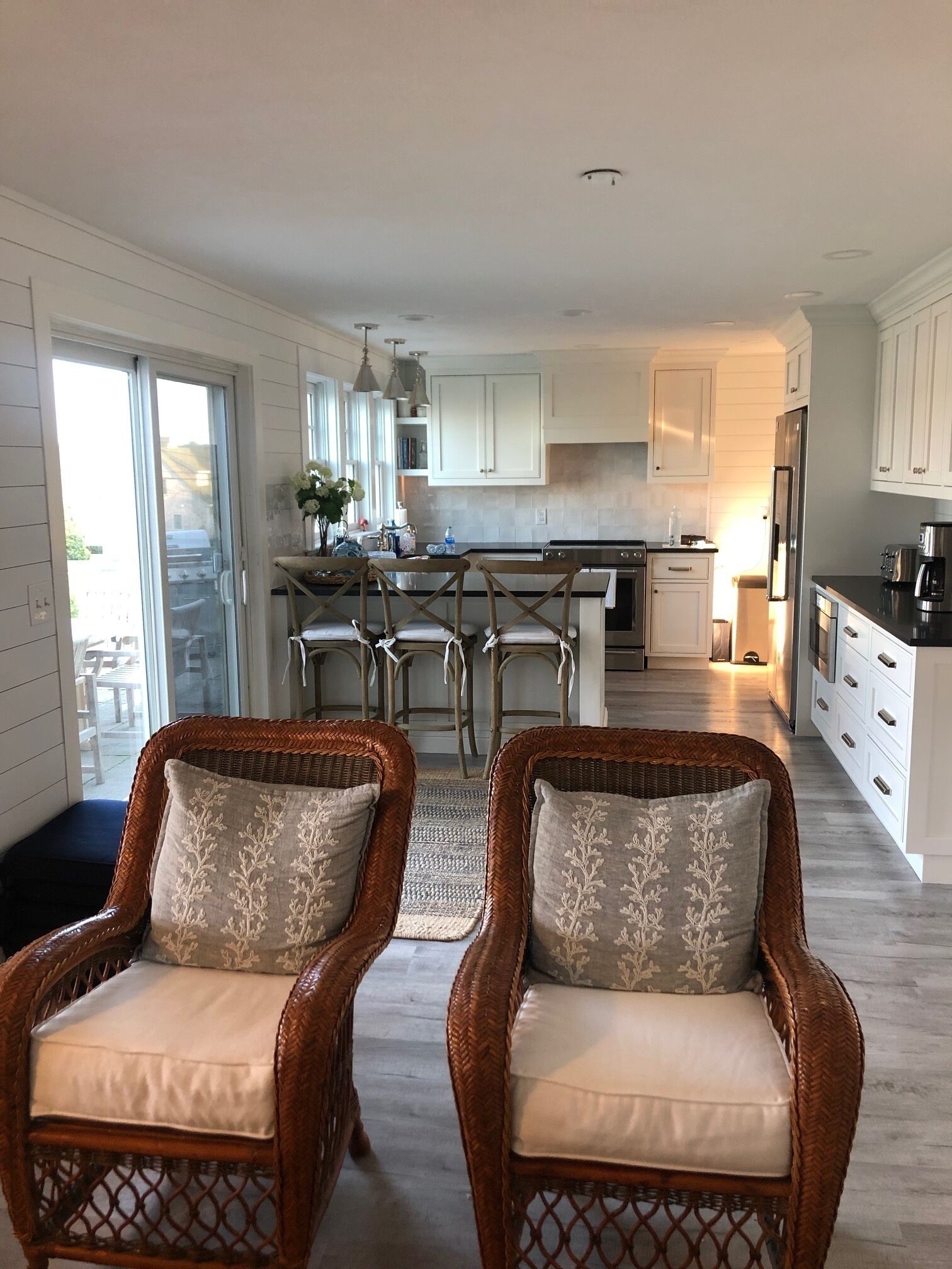 A modern kitchen with white cabinets, a central island with bar stools, and wicker chairs with cushions.