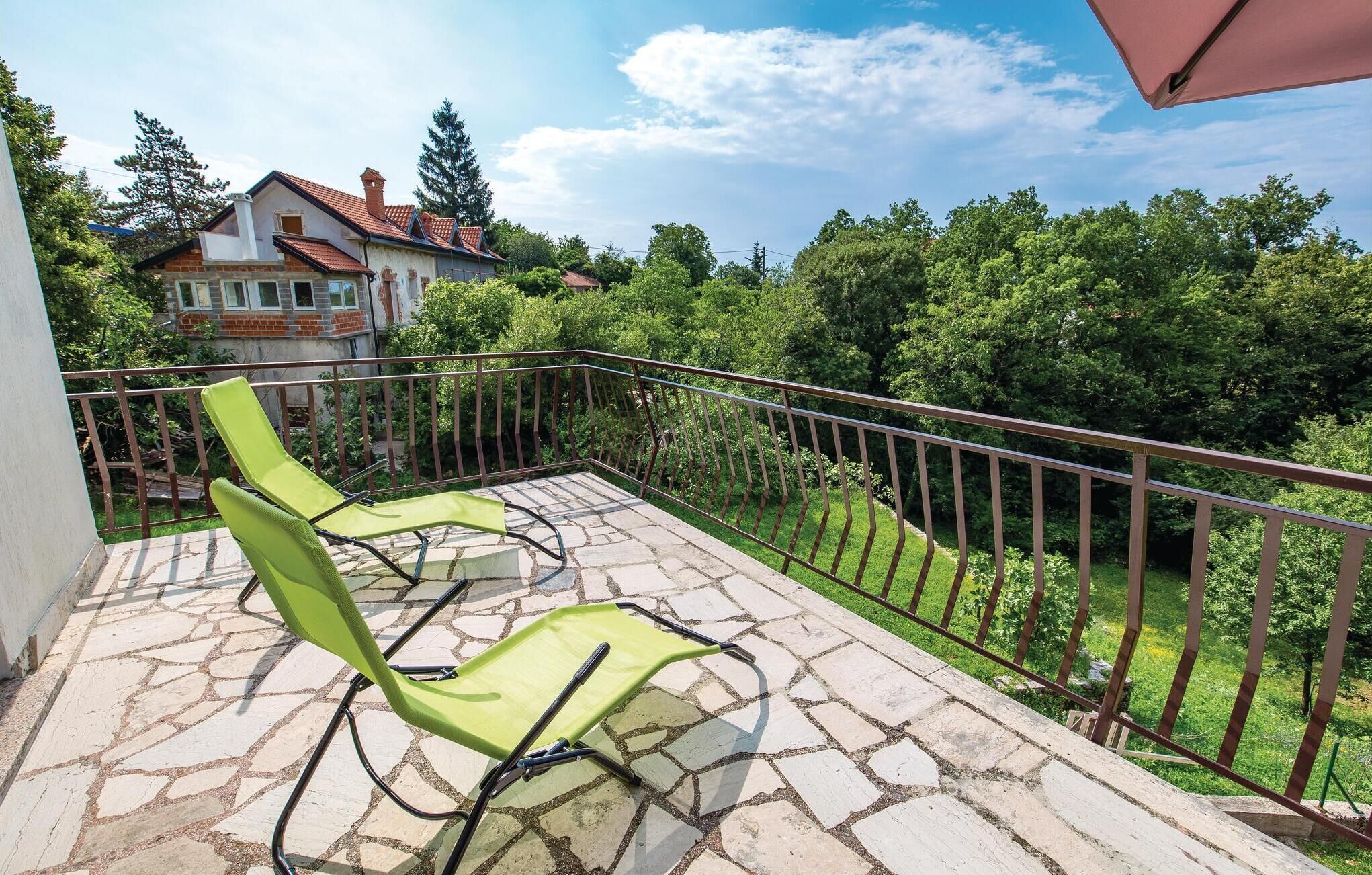 A balcony with two green chairs, a stone-tiled floor, and a view of a house and trees.