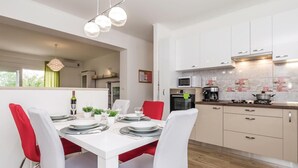 Een moderne keuken met witte kasten, een magnetron, een koffiezetapparaat en een fornuis. Er is een eethoek met een witte tafel en rode stoelen.