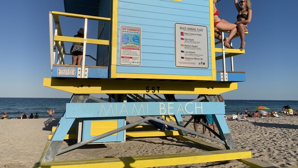 Uma torre de salva-vidas em Miami Beach, com esquema de cores azul e amarelo, uma placa indicando bandeiras de alerta para a praia, e o nome 'MIAMI BEACH' na estrutura.