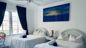 Quarto de hotel com duas camas, um ventilador de teto, uma pintura de pôr do sol e vista para edifícios pela janela.