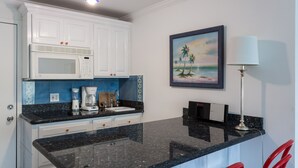 Cozinha com armários brancos, bancada preta, micro-ondas, cafeteira e um quadro com uma paisagem de praia.
